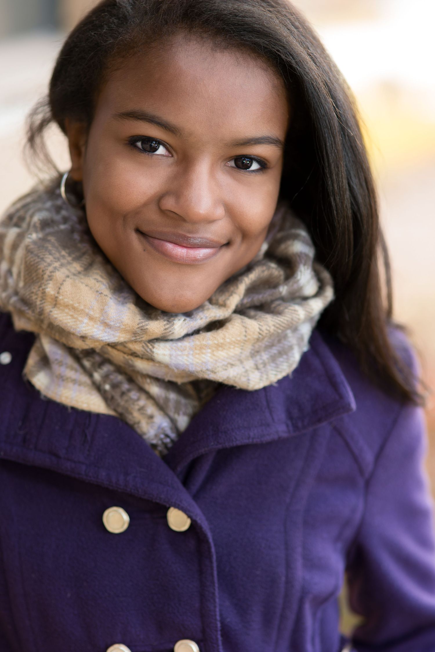 Young Black woman smiling warmly, wearing a plaid scarf and purple coat, photographed outdoors in natural light.