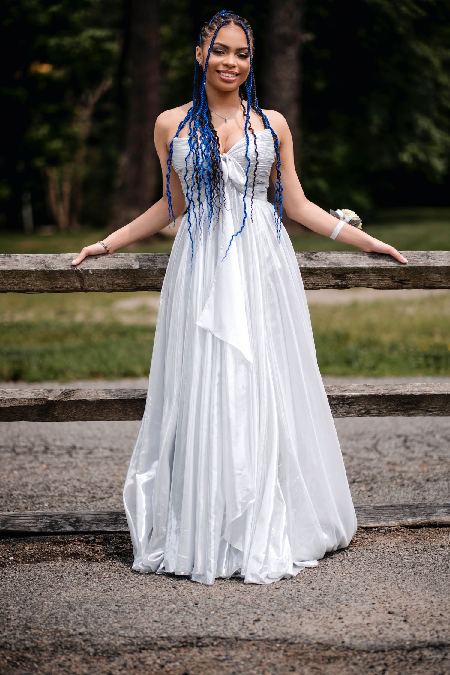 Young woman in white halter gown with blue braids poses gracefully outdoors near a rustic wooden fence.