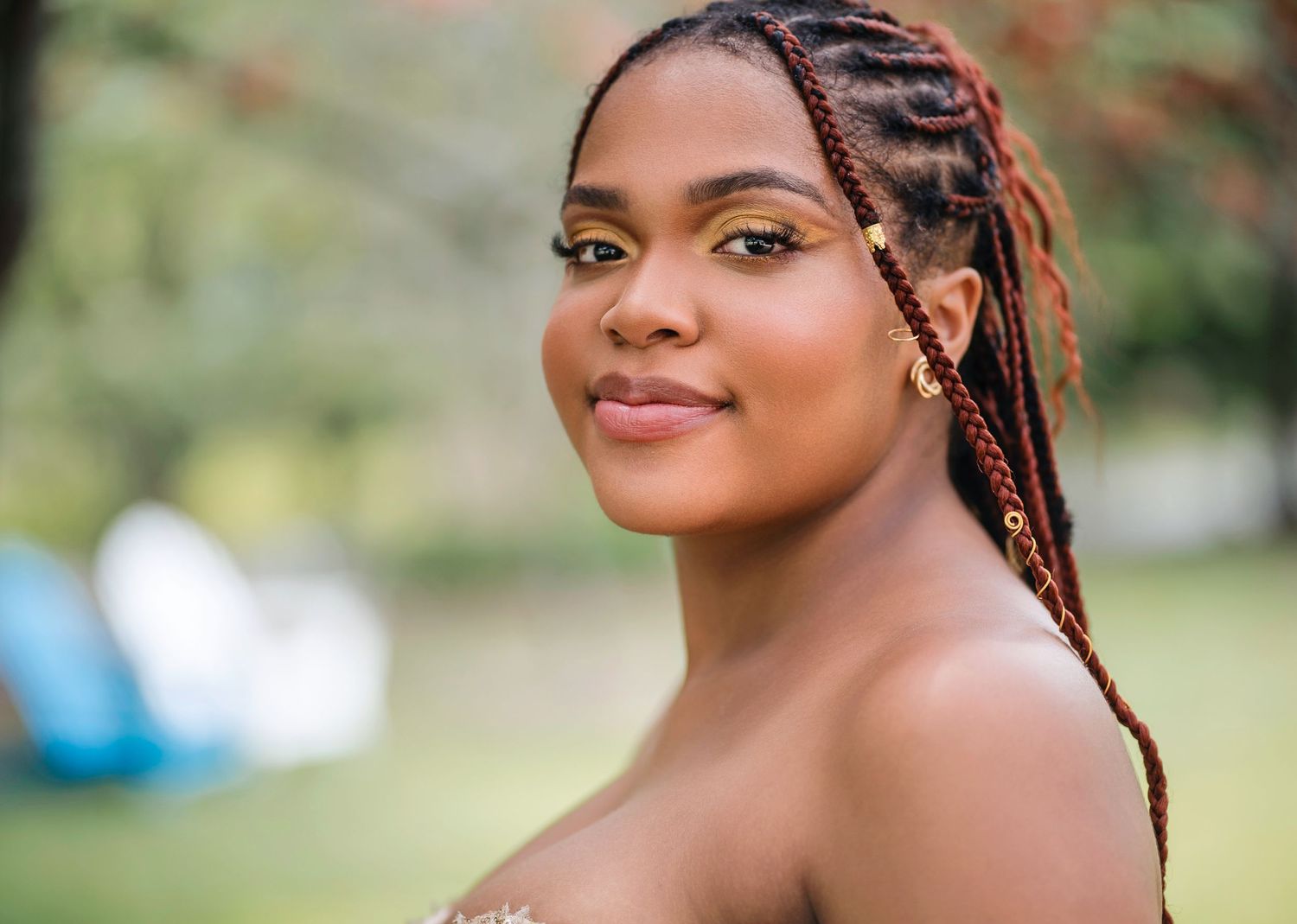 Young woman with braided hair and gold eye makeup smiling outdoors in a lush green park setting.