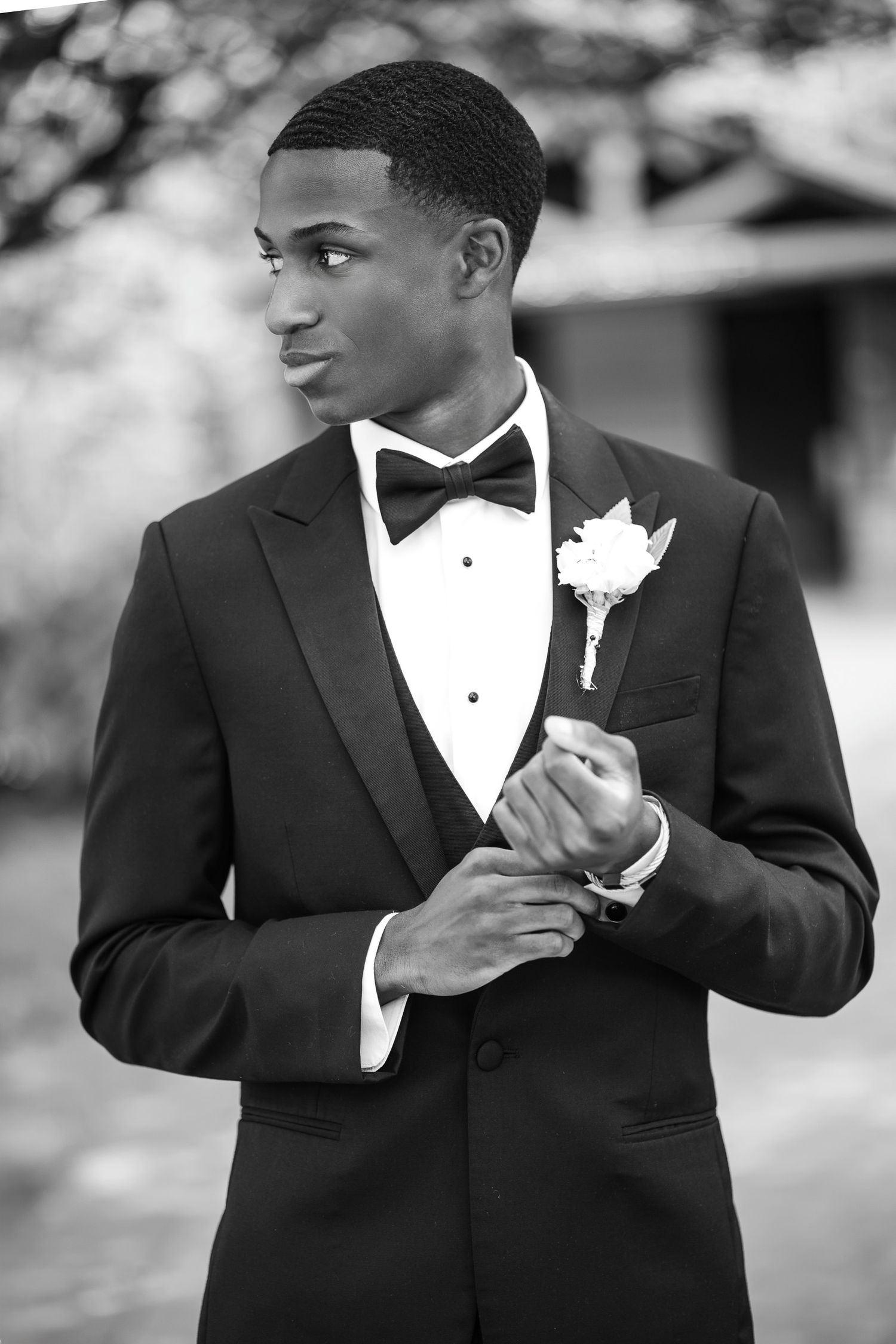 Young groom in black tuxedo with bow tie and boutonniere adjusting cufflinks outdoors in black and white photo.