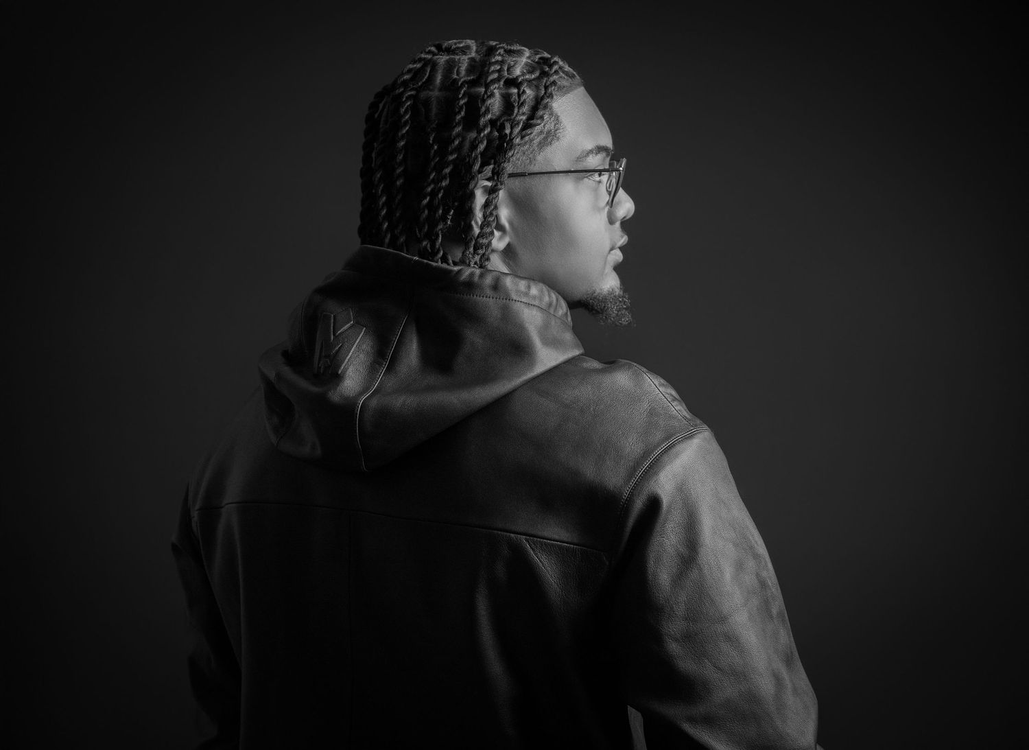 Black and white portrait of a person with braids and glasses wearing a leather jacket, photographed from behind.