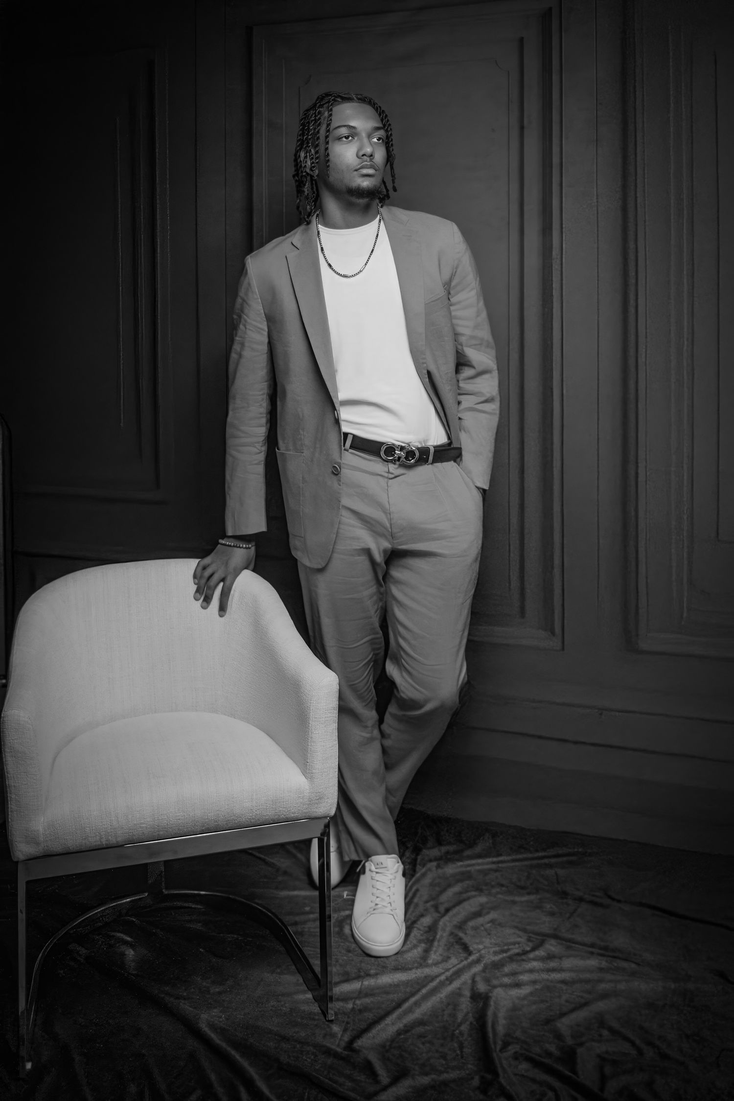 Black and white portrait of a stylish young man in a suit leaning against a chair in an elegant room.