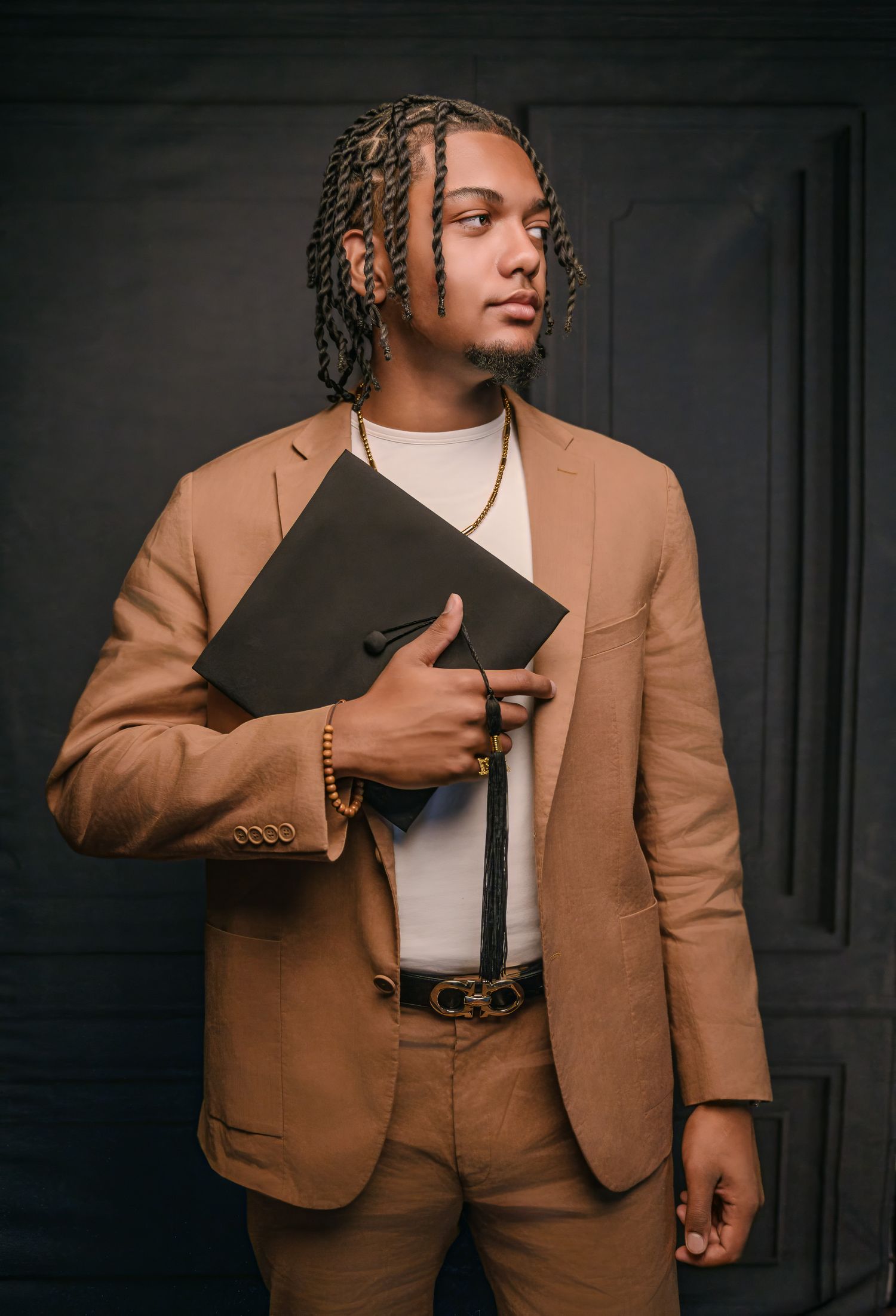 Young man in tan suit holding graduation cap, wearing gold jewelry and braided hair, posed against dark background.