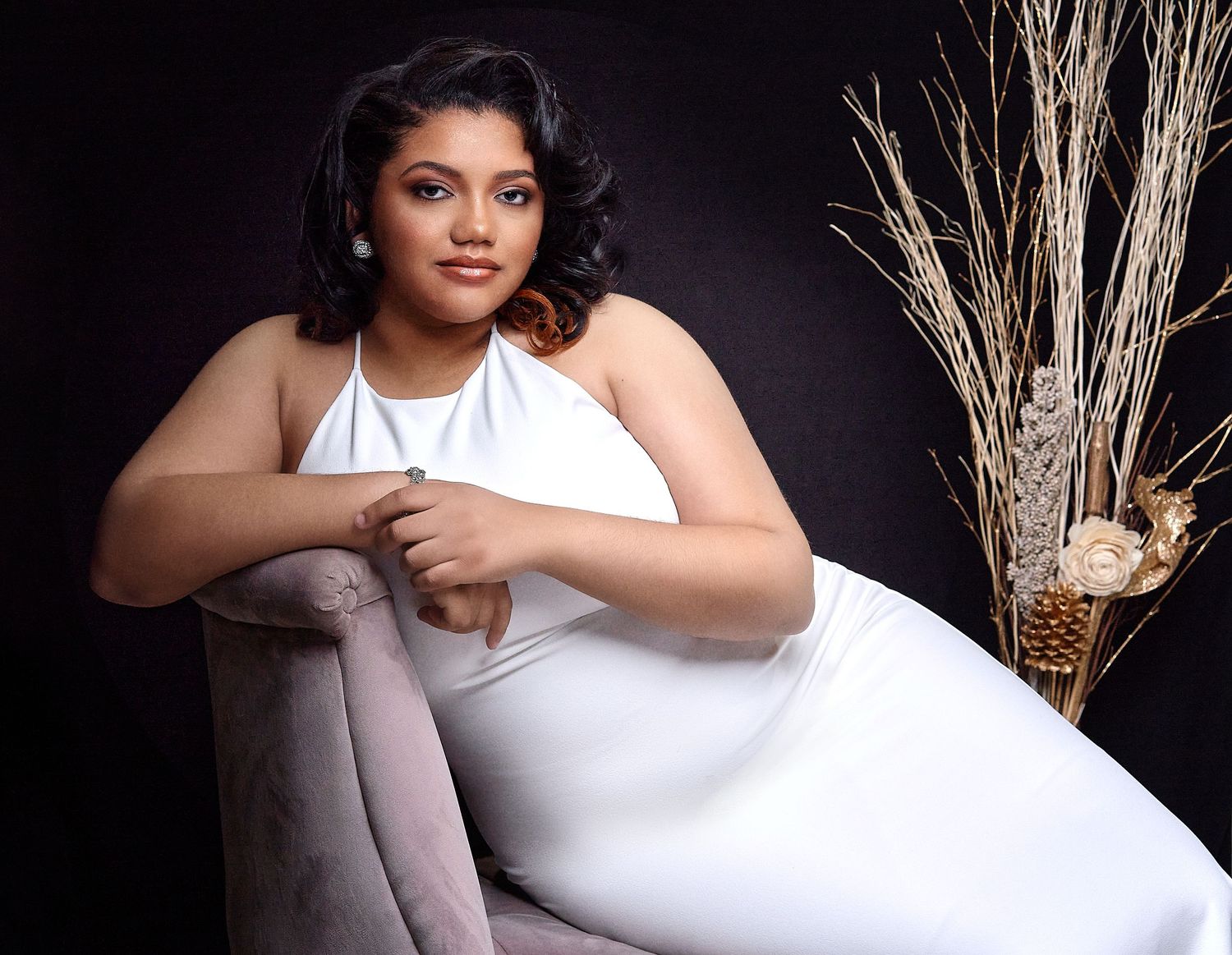Young woman in white halter dress poses elegantly in studio portrait with decorative branches in background.