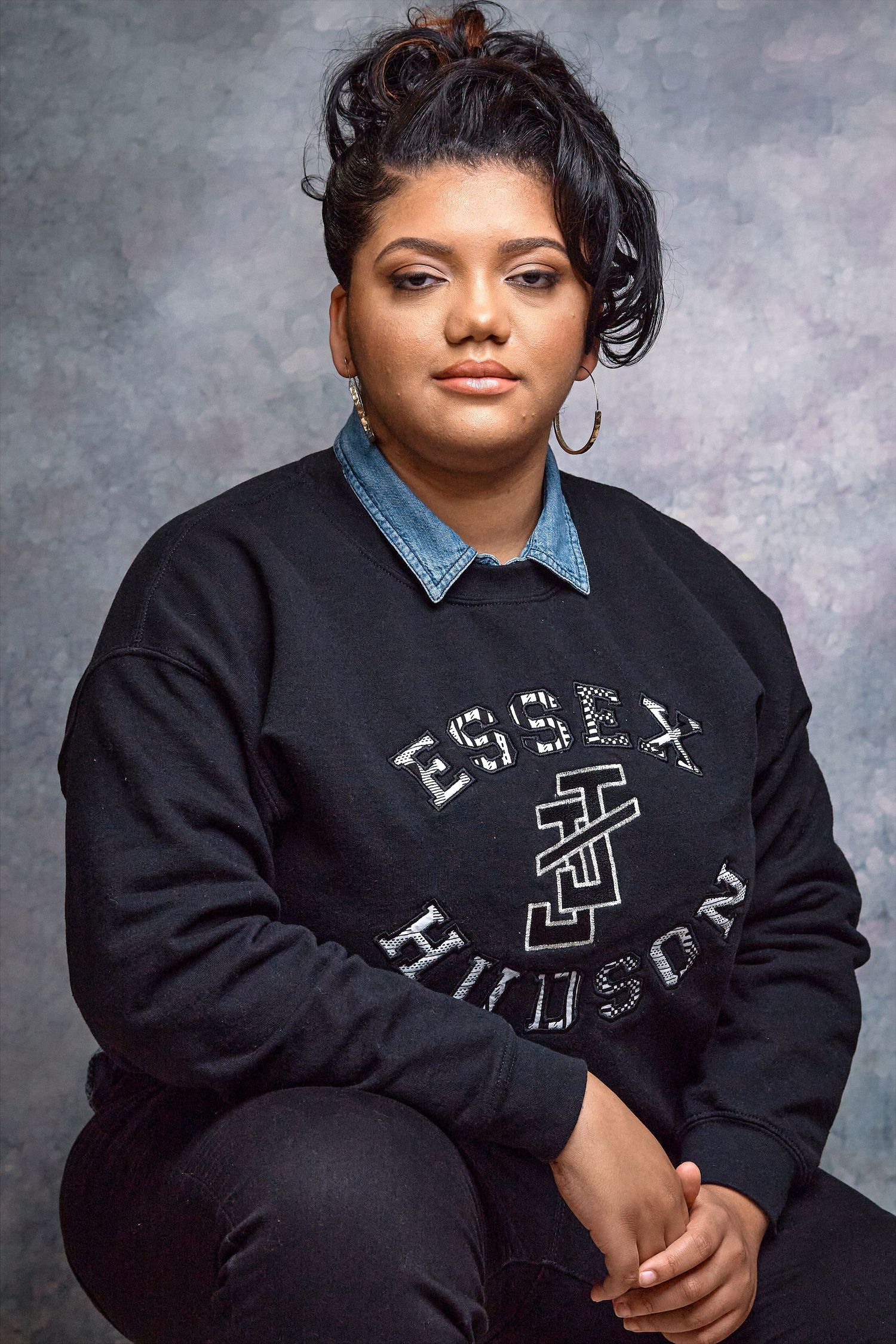 Woman wearing black Essex Hudson graphic sweatshirt over denim shirt, dark hair up, hoop earrings, studio portrait.