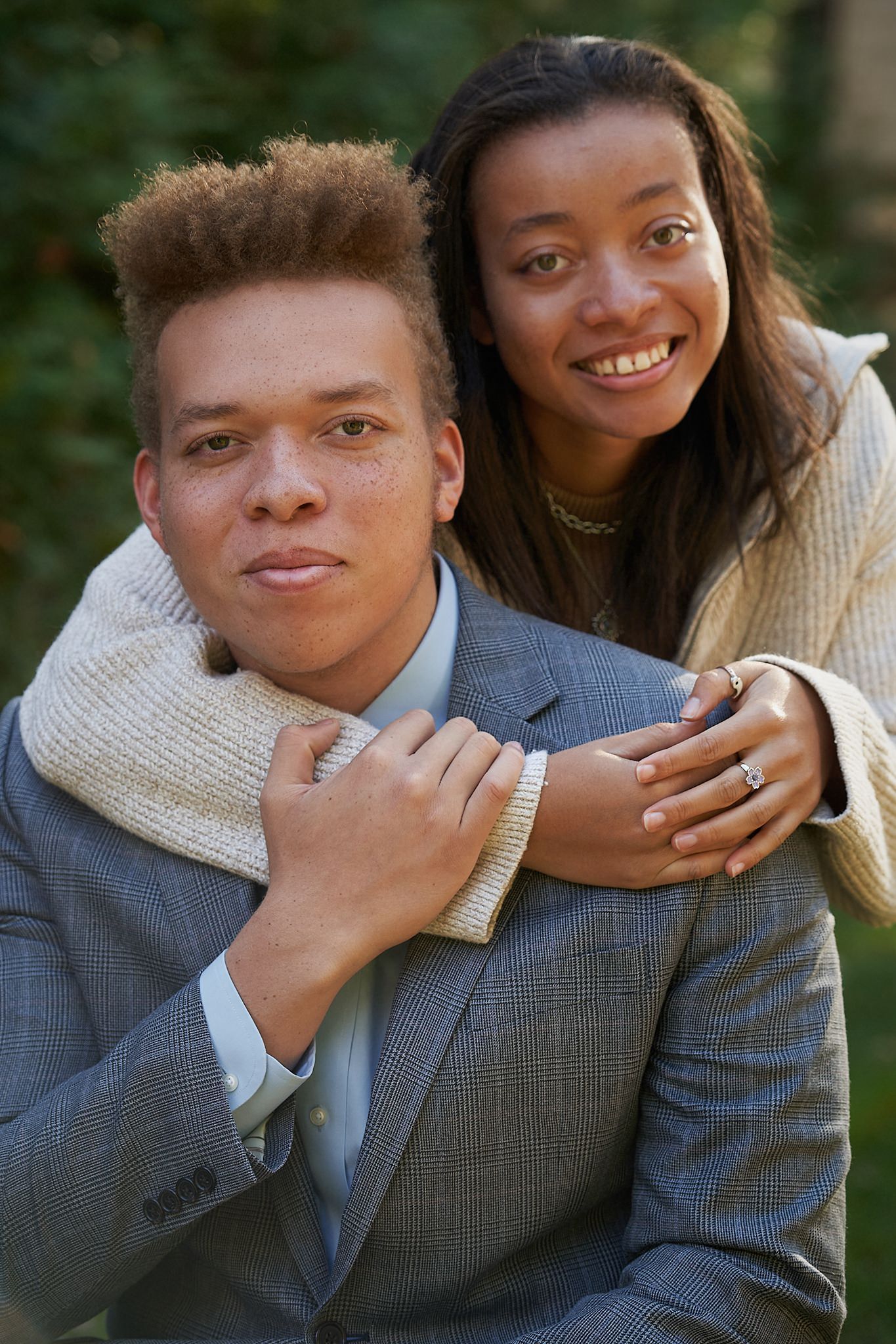 A young couple poses outdoors, woman smiling and embracing man in gray suit from behind, surrounded by greenery.