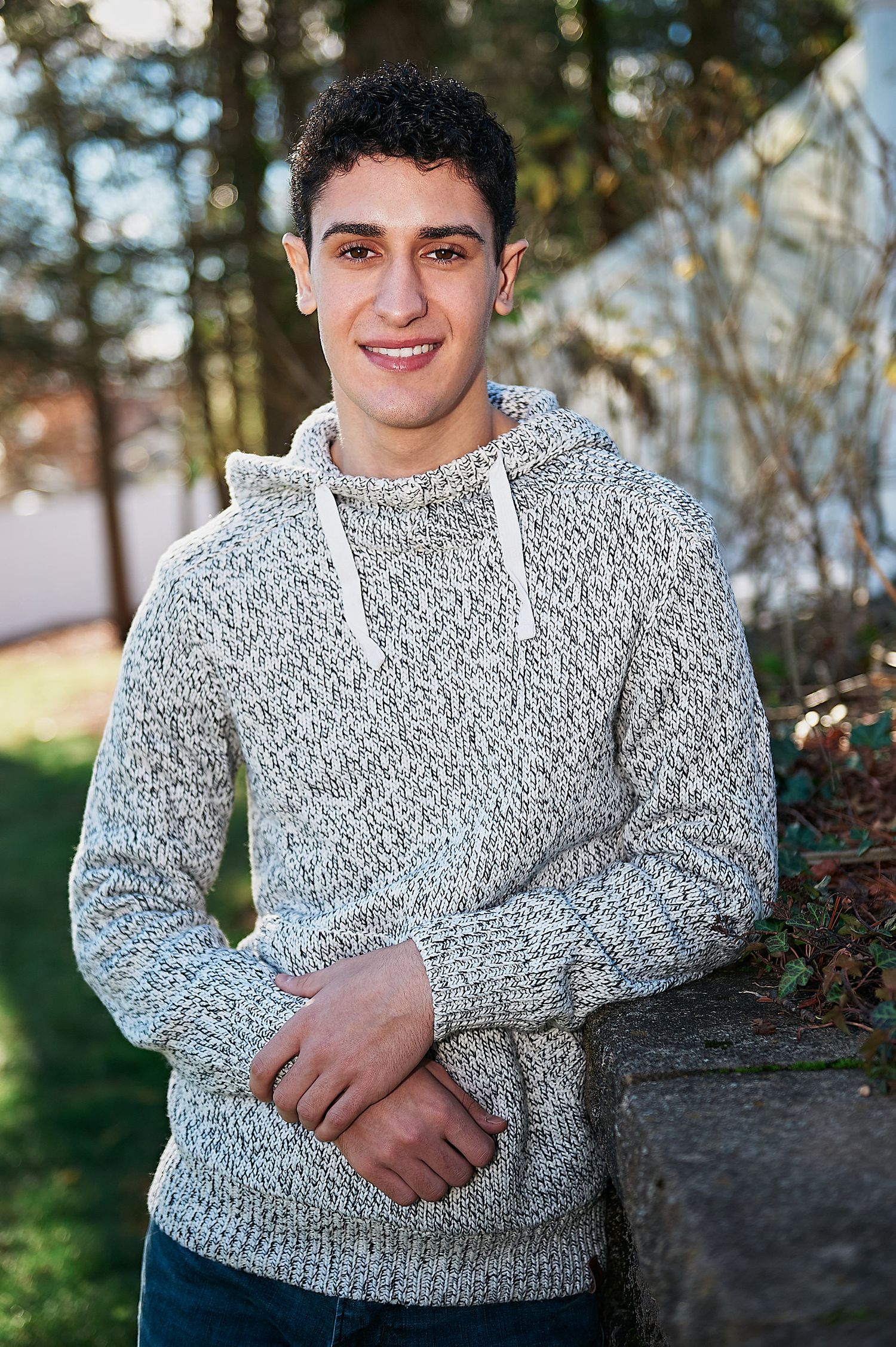 Young man smiling in a gray marled hoodie sweater, posing outdoors near a stone wall with trees in background.