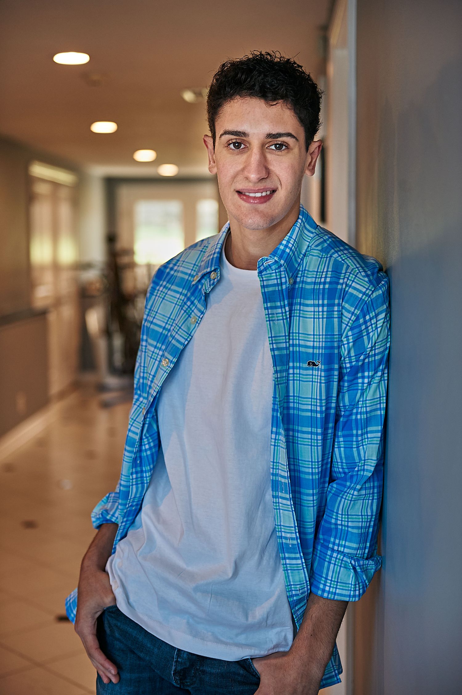 Young man in blue plaid shirt and white tee smiles leaning against wall in bright modern hallway.