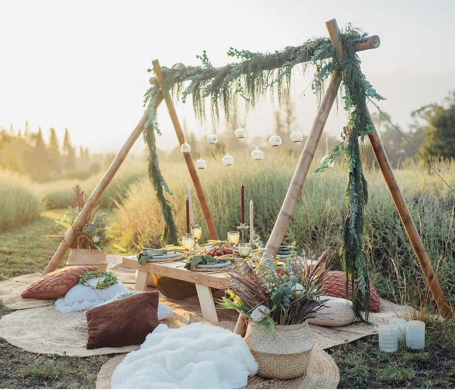 Boho-style picnic setup with wooden arch, cushions, and greenery decorations in an outdoor field at sunset on Maui, Hawaii.