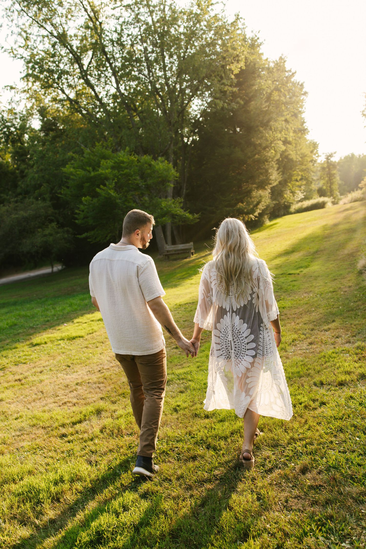 Couple walks hand in hand through sunlit grassy hill during golden hour.