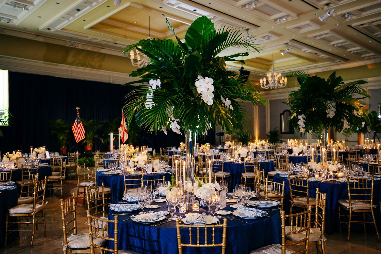 Elegant ballroom tables decorated with blue linens, golden chairs, and tall tropical centerpieces for luxury wedding reception.
