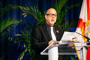 Speaker in black suit and glasses presents from clear podium with palm plants and flags in background.