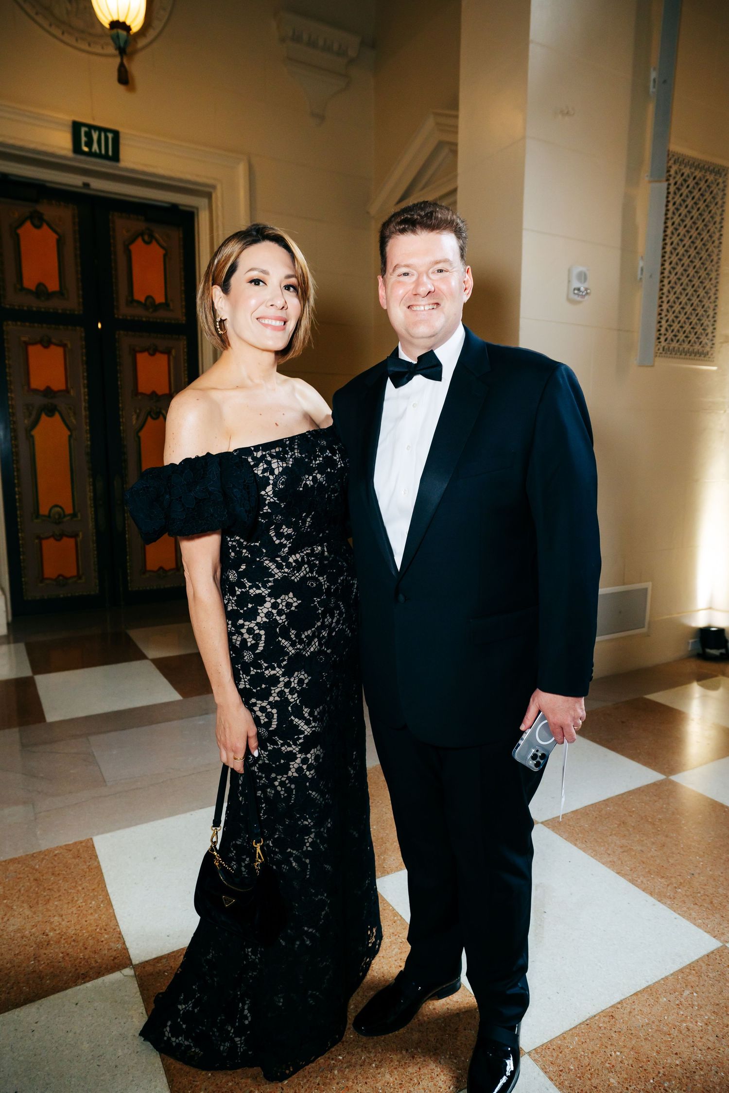Elegant couple in formal attire attending a black-tie event with an off-shoulder black lace gown and classic tuxedo.