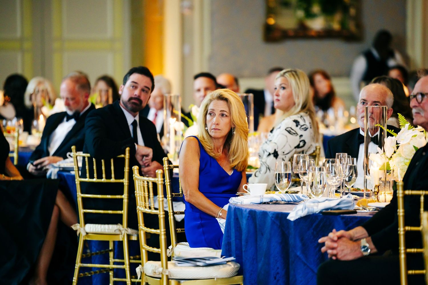 Wedding guests enjoy an elegant dinner reception with blue decor and gold chiavari chairs at round tables.