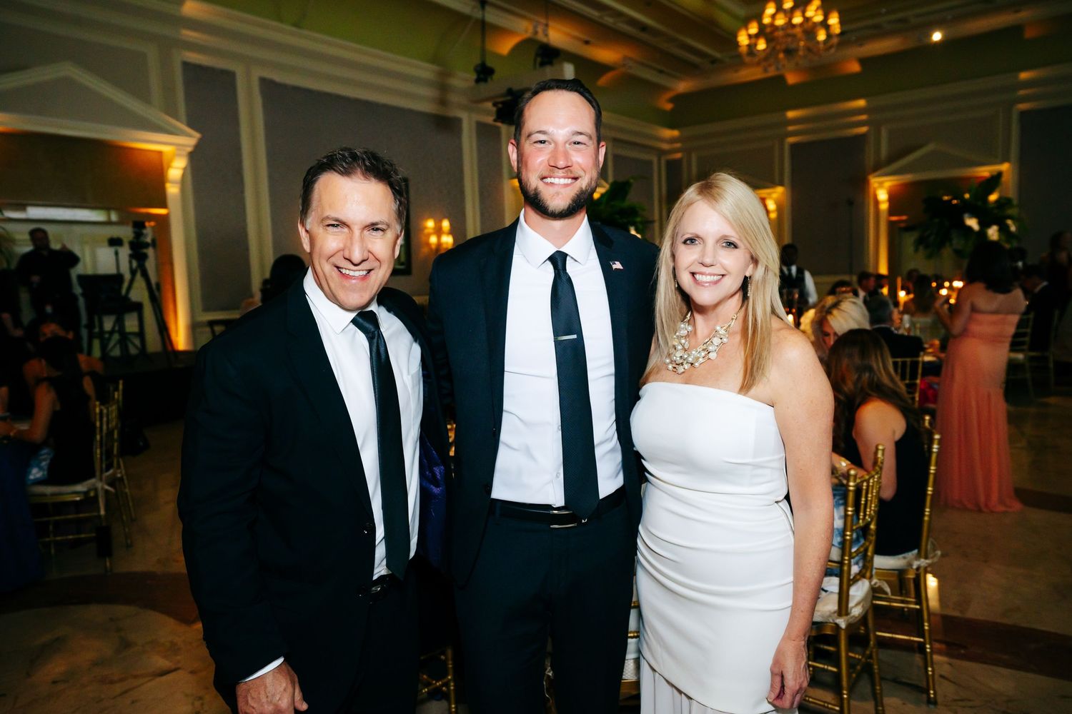 Three guests in formal attire smile together at an elegant event with crystal chandeliers.