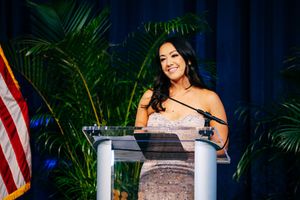 Event speaker presenting at an elegant podium with palm decorations and an American flag in the background.