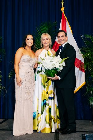 Formal group photo in front of a blue curtain and Florida state flag featuring elegant evening attire and floral bouquet.