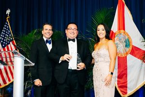 Three attendees in formal attire pose at an event with American and Florida state flags as backdrop.