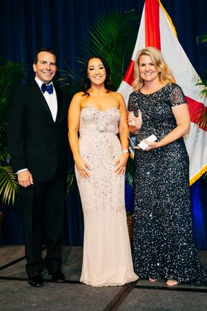 Three people pose formally in evening wear against blue curtain backdrop with state flag.