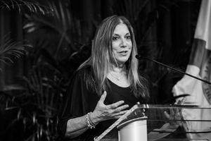 A speaker presents at a podium against a backdrop of tropical plants in black and white photography.