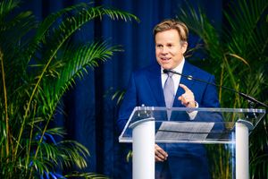 Speaker presents at a podium with tropical palm plants decorating the blue backdrop.