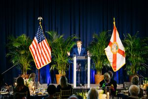 Speaker at podium with American and Florida state flags flanked by palm plants on stage.
