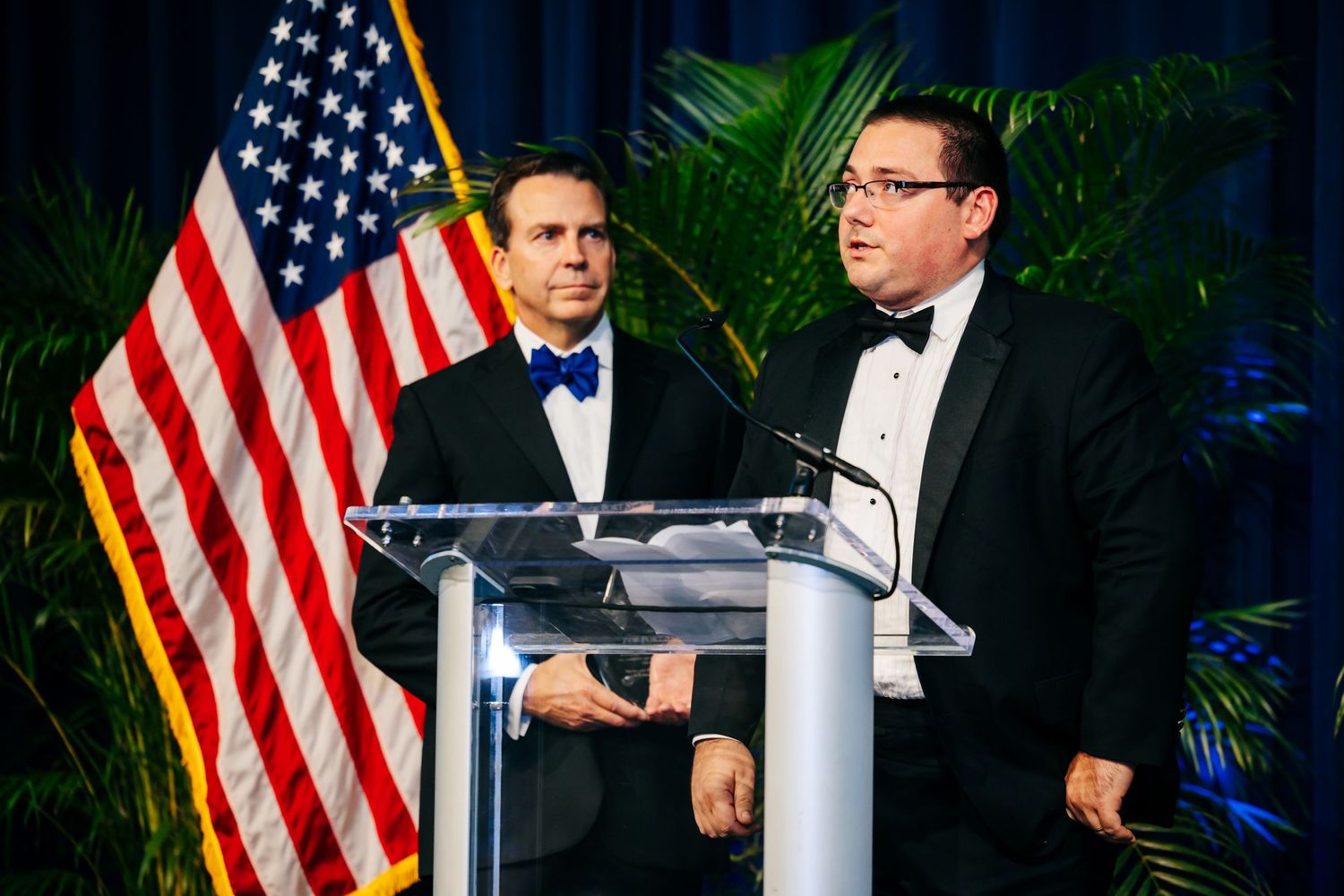 Two speakers stand at a podium in formal attire with an American flag and tropical plants in the background.