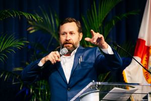 A speaker in navy suit gestures while presenting at podium with palm plants in background.