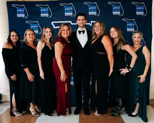Large group photo of elegantly dressed guests in black formal attire posing against a dark blue backdrop.