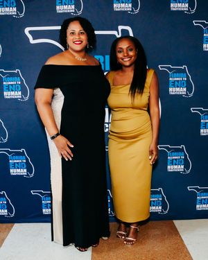 Two attendees pose at an event against a blue backdrop with repeated logo patterns.