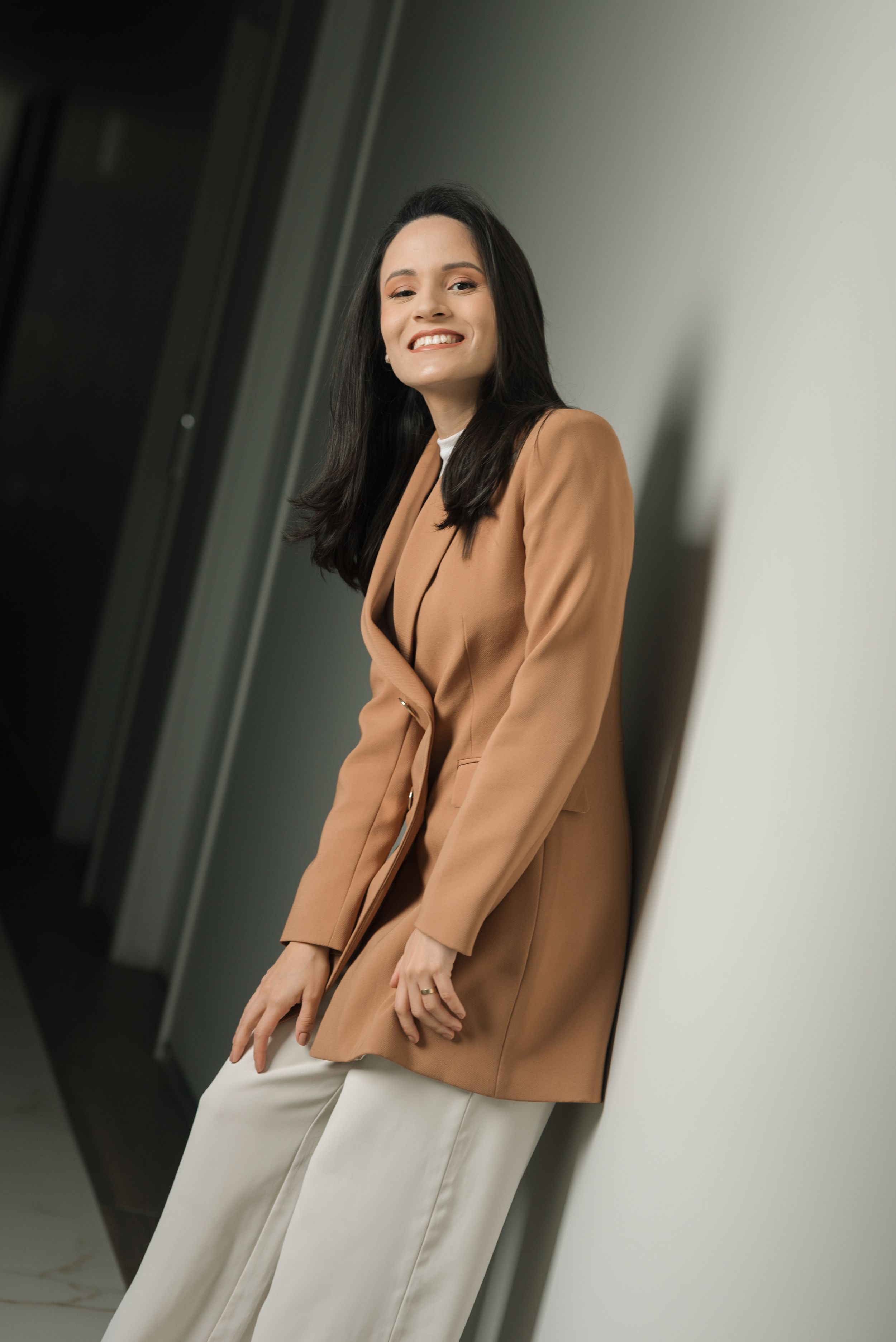 Business professional in tan blazer and white pants standing in modern office hallway.