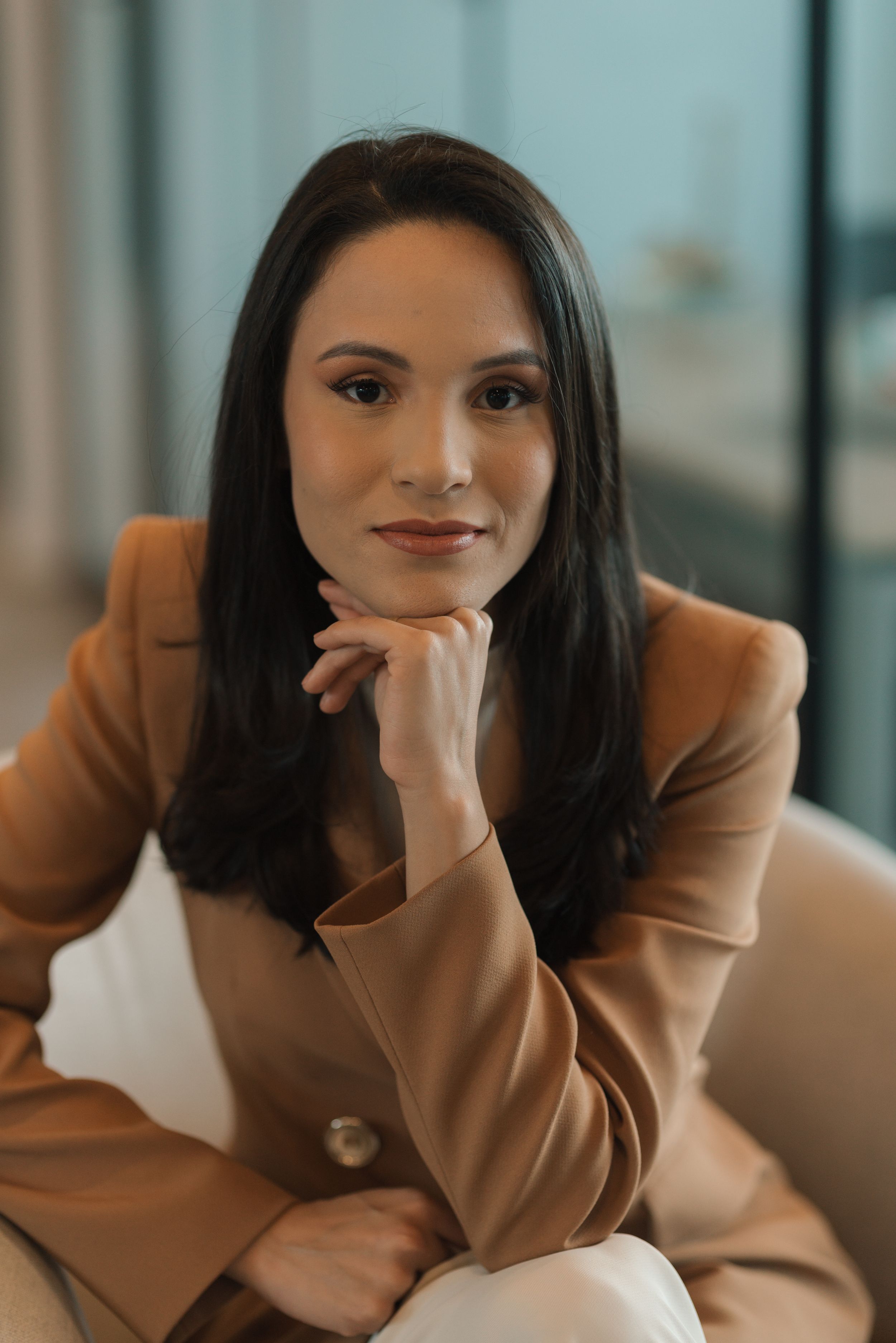 Professional headshot of person in tan blazer with hand under chin in modern office setting.