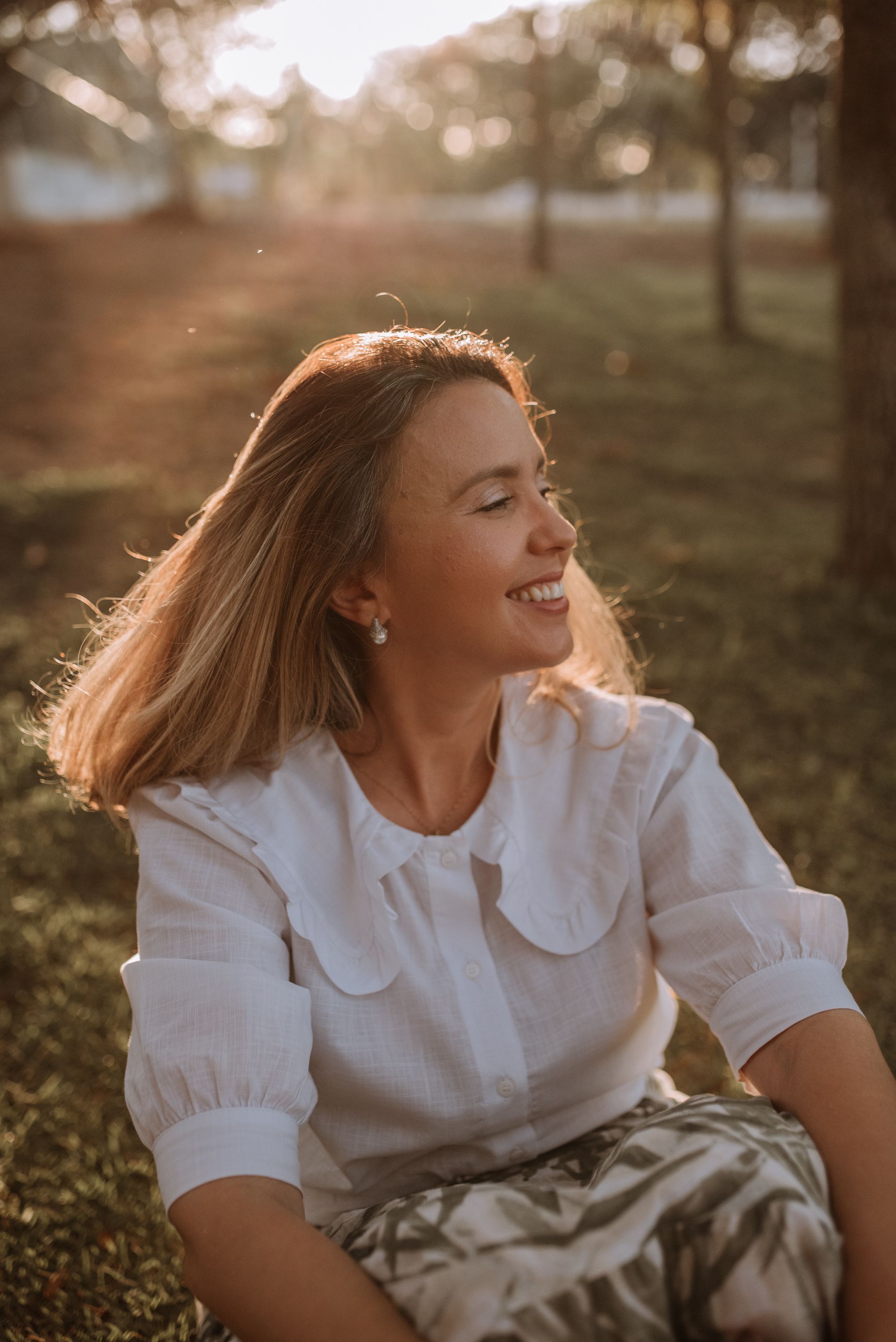 Sunset portrait of a person in white blouse smiling while sitting on grass.