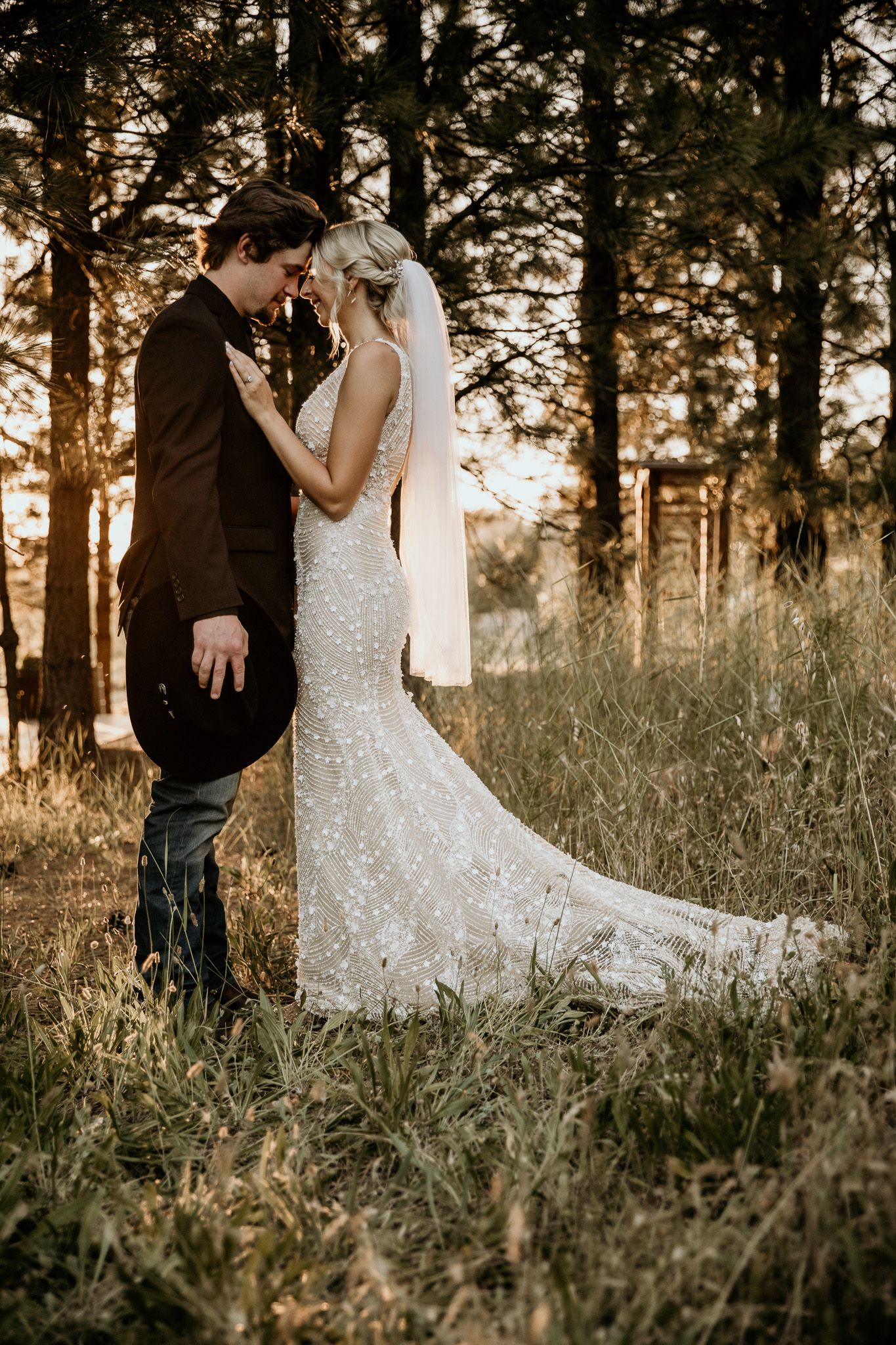 A romantic sunset wedding photo of a couple standing in a pine forest with a flowing white mermaid gown.