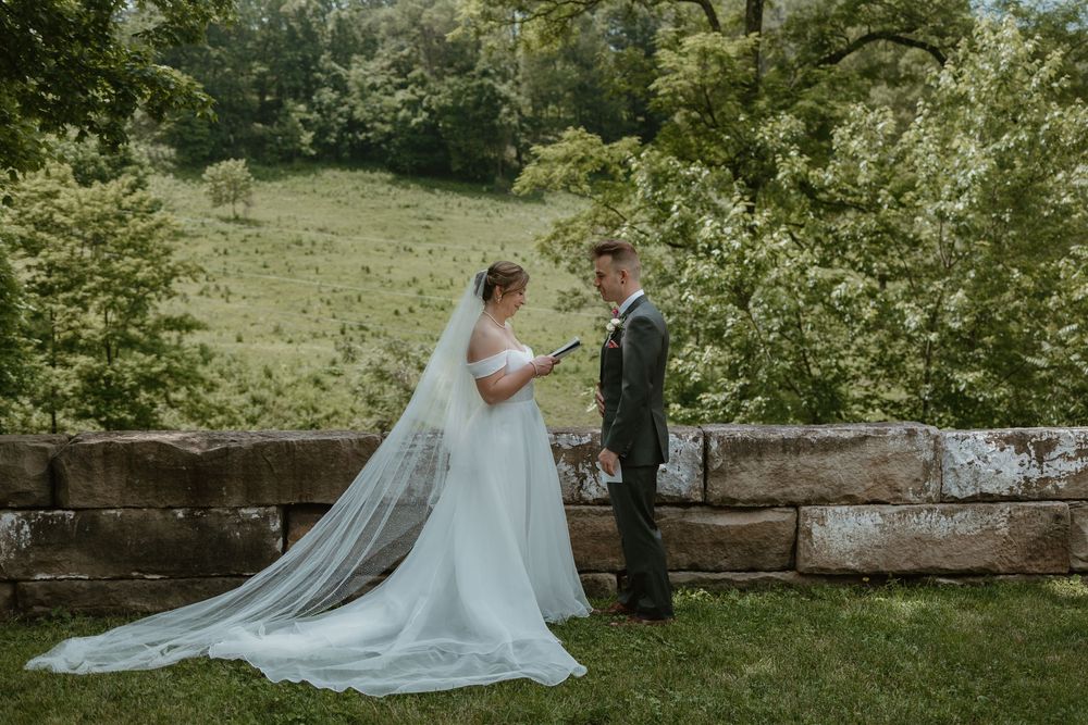 Bride in flowing veil reads vows to groom outdoors near stone wall surrounded by green hills.