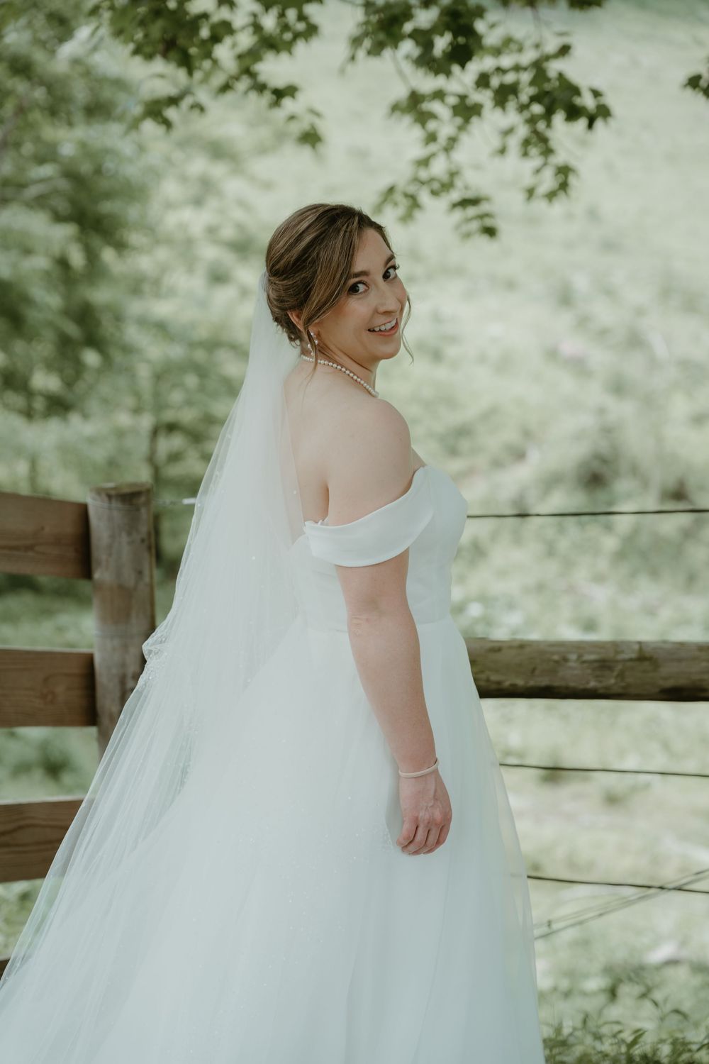 Bride in off-shoulder white gown with veil smiles over shoulder near rustic wooden fence.