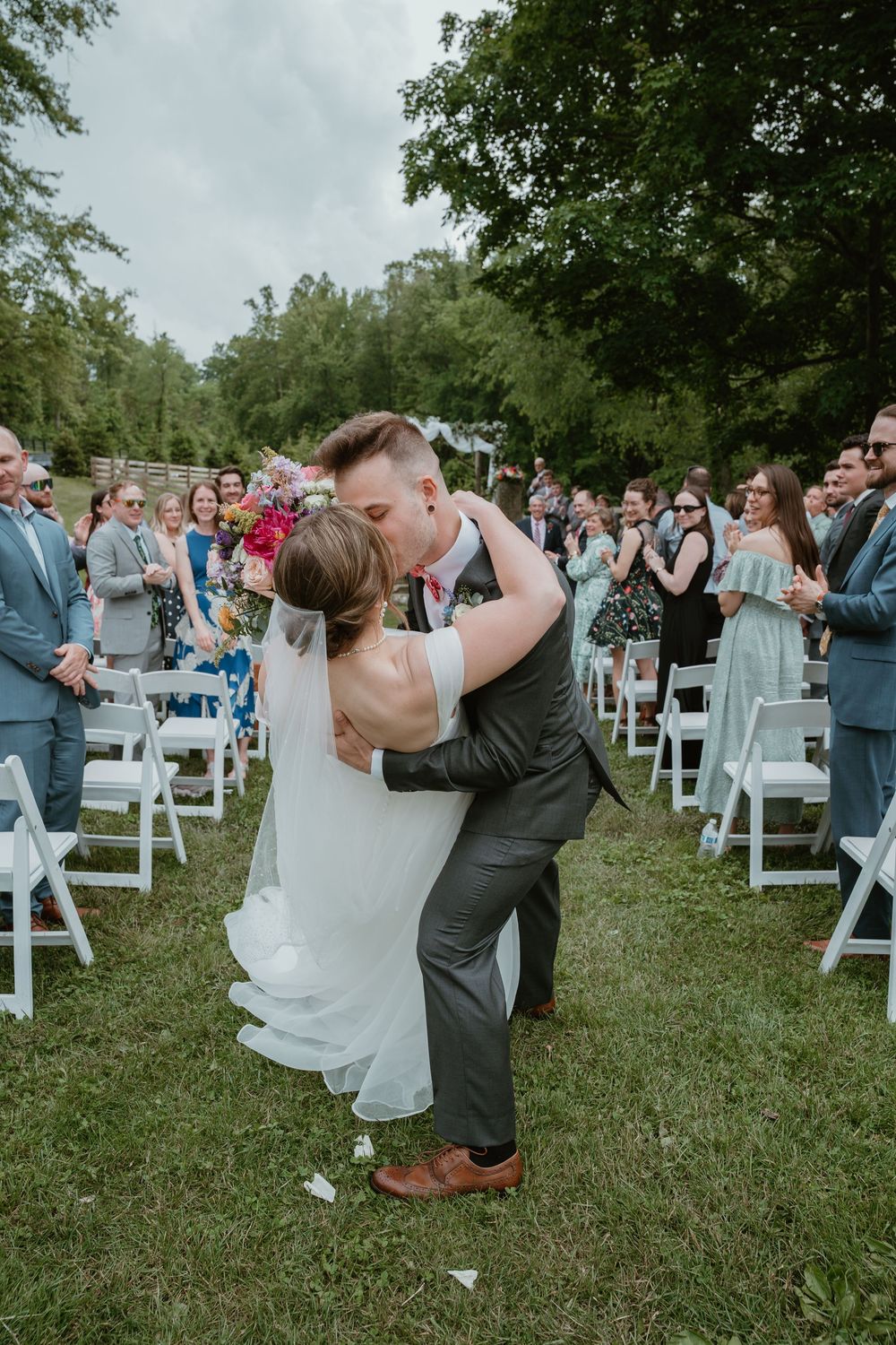 Groom lifts bride for a kiss at outdoor wedding ceremony as guests cheer and celebrate.