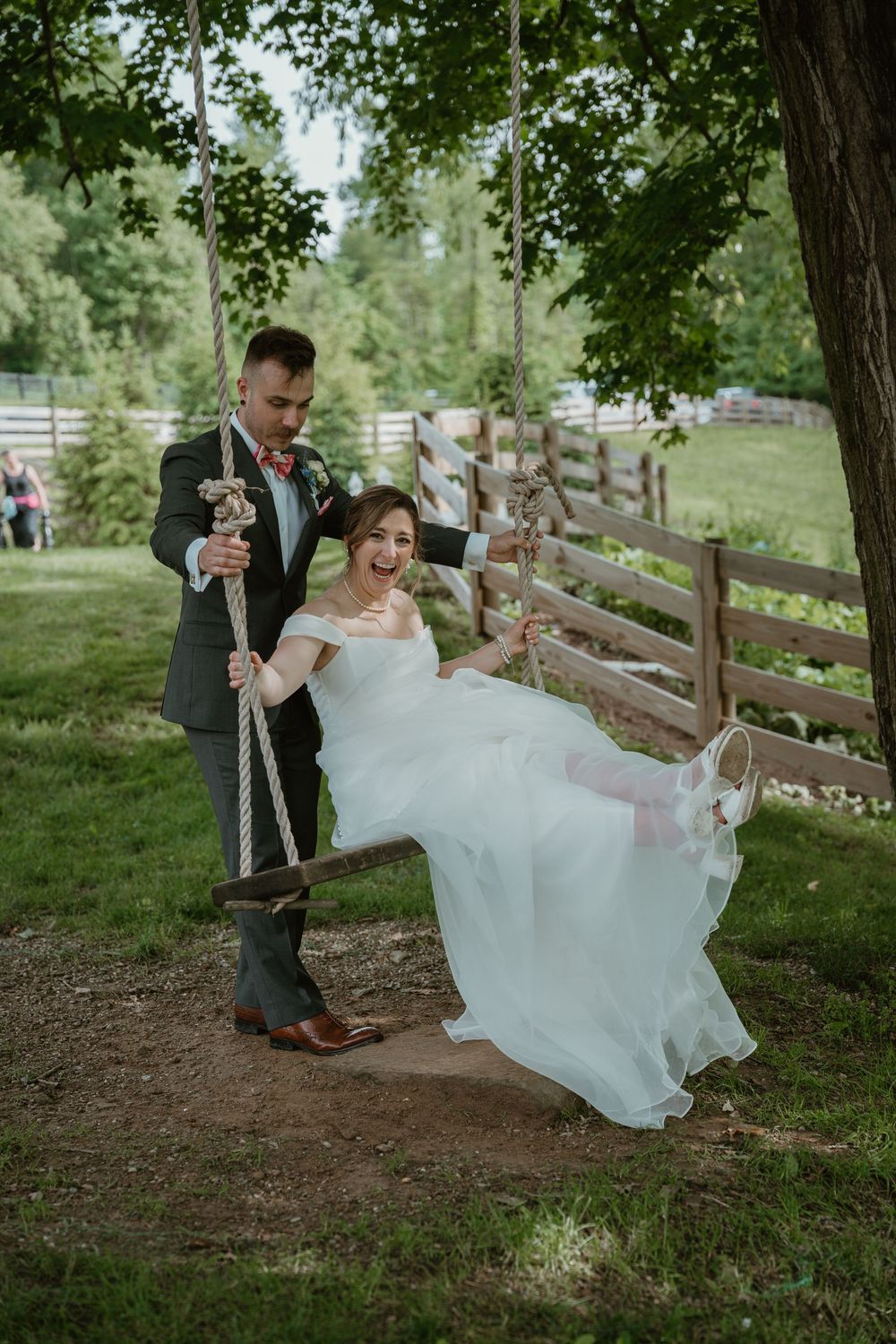 Groom pushes laughing bride on rope swing at outdoor rustic farm wedding venue.