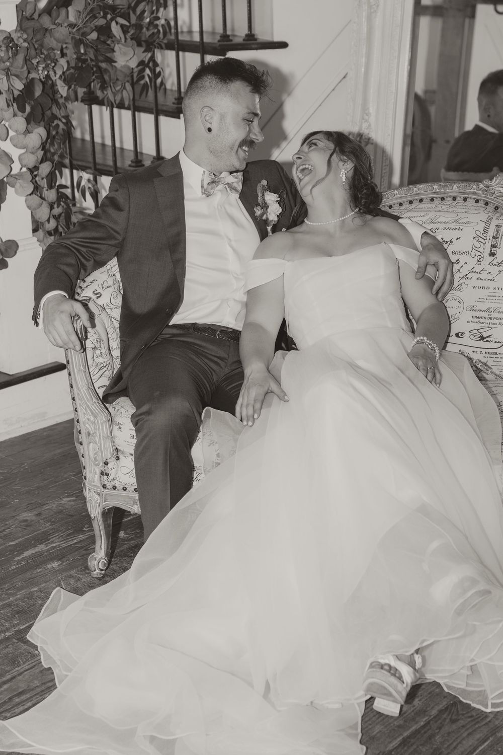 Bride and groom laughing together on ornate vintage sofa in black and white wedding portrait.