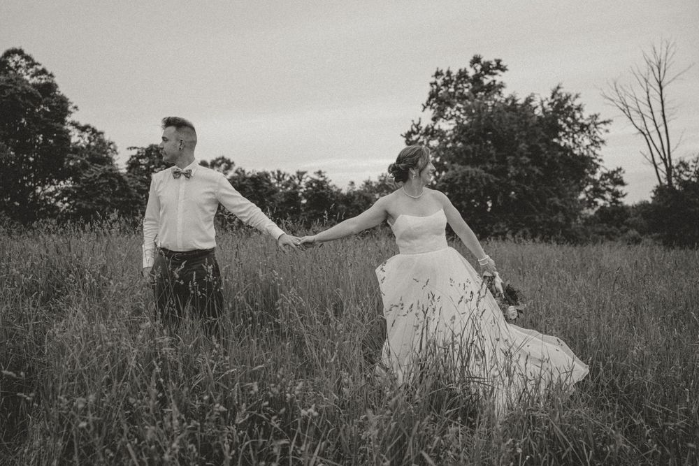 Black and white photo of bride and groom holding hands walking through tall grass field.