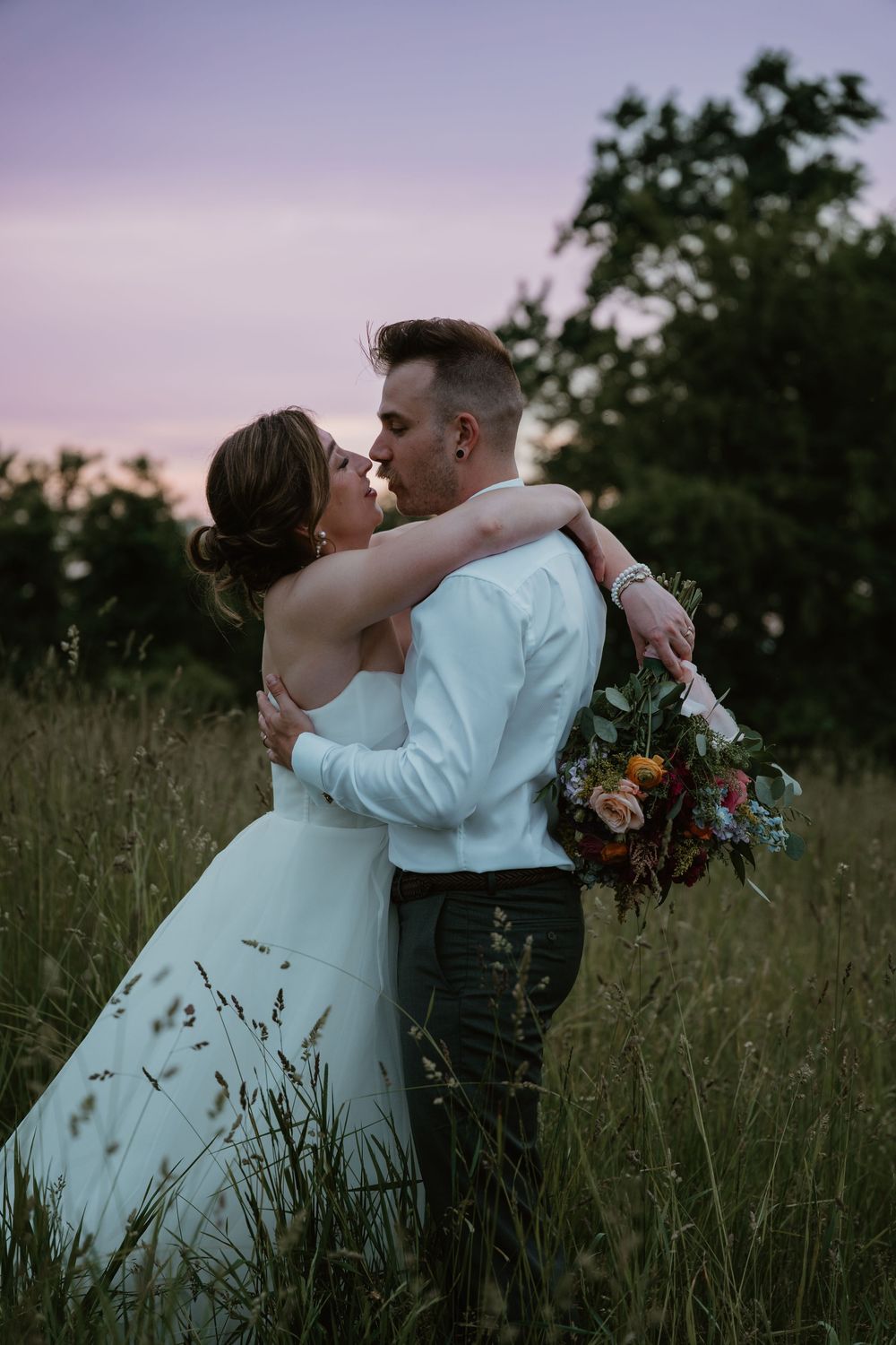 Bride and groom embracing and kissing at sunset in a lush green meadow, bride holding colorful bouquet.