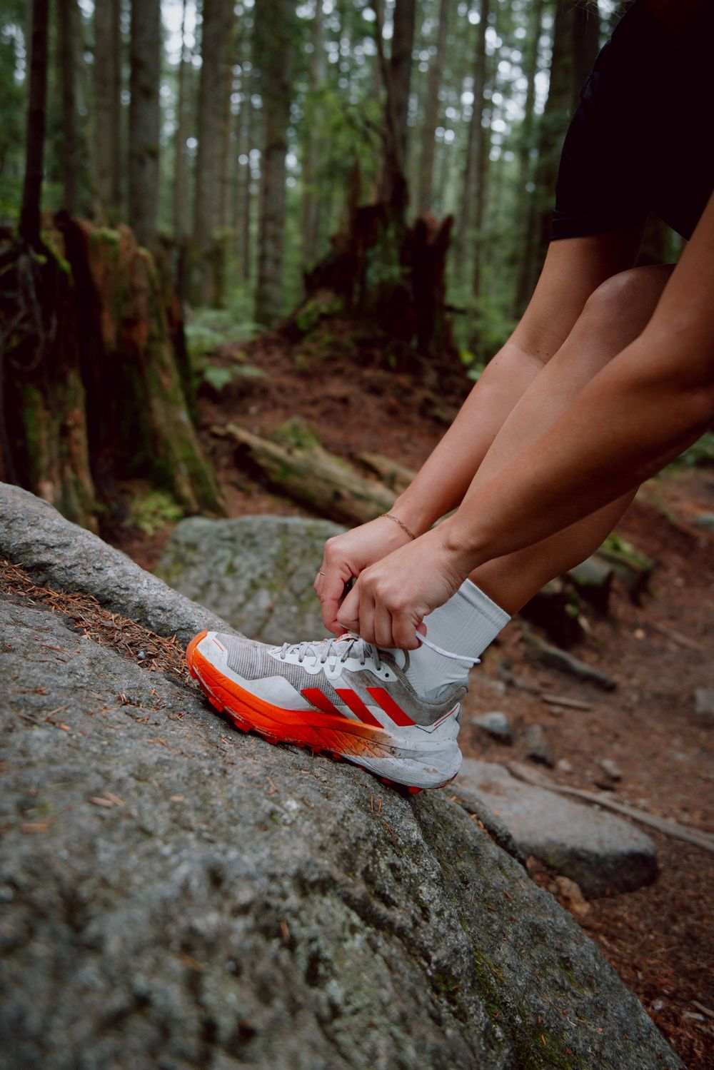 Close-up shot of someone lacing up grey and orange athletic shoes while sitting on a rock in a forest setting.