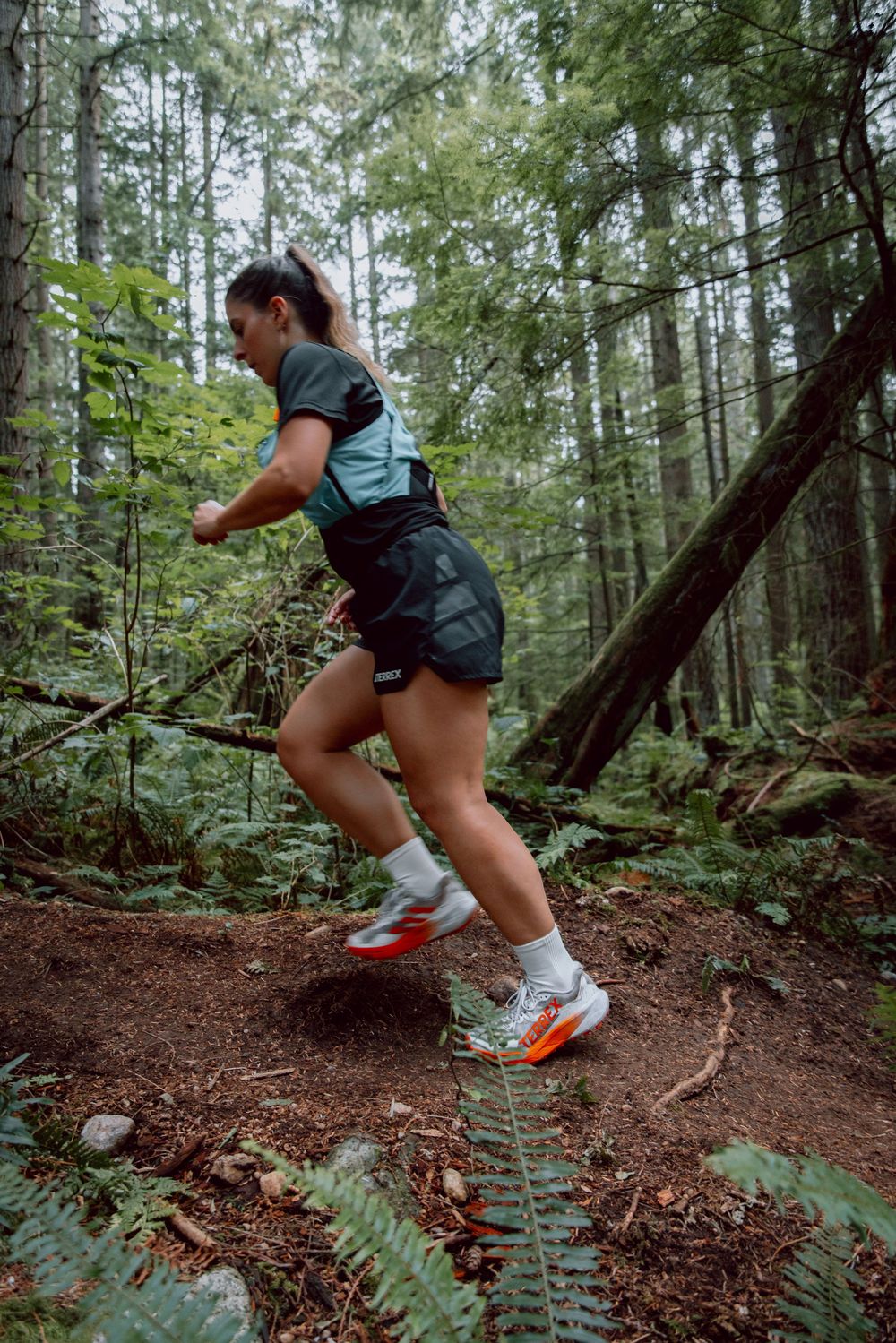 Trail runner in workout gear and orange sneakers navigating a forest path surrounded by lush greenery and tall trees.