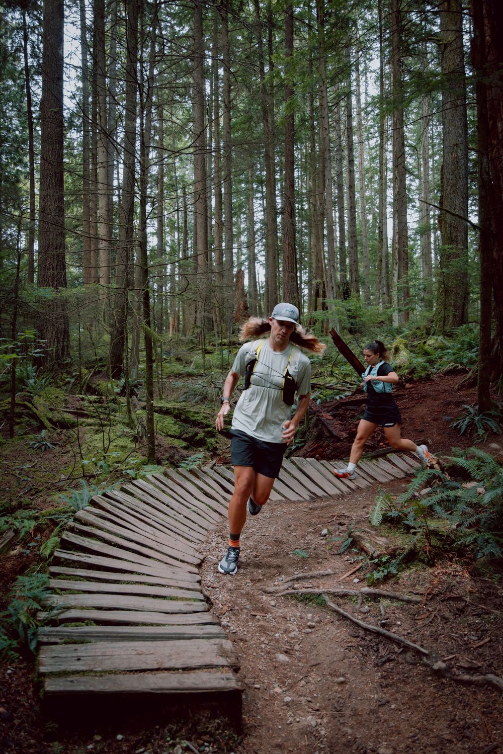A mountain biker navigates a winding wooden berm trail through a dense forest of tall evergreen trees.