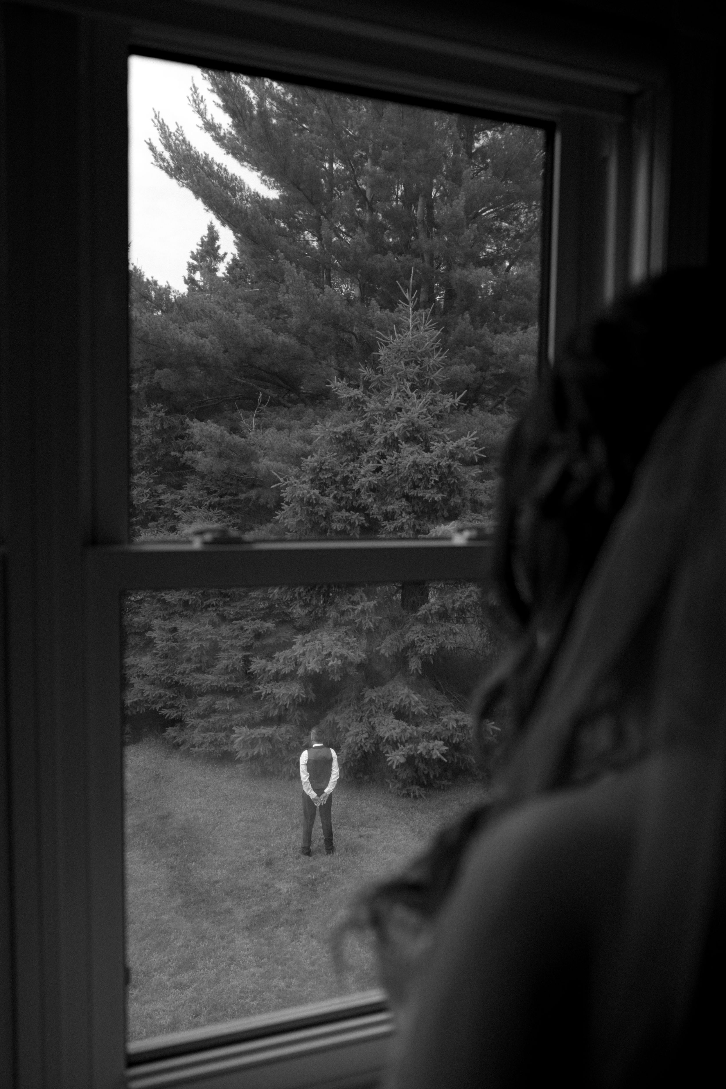Black and white view through window showing solitary figure standing in yard with evergreen trees in background.