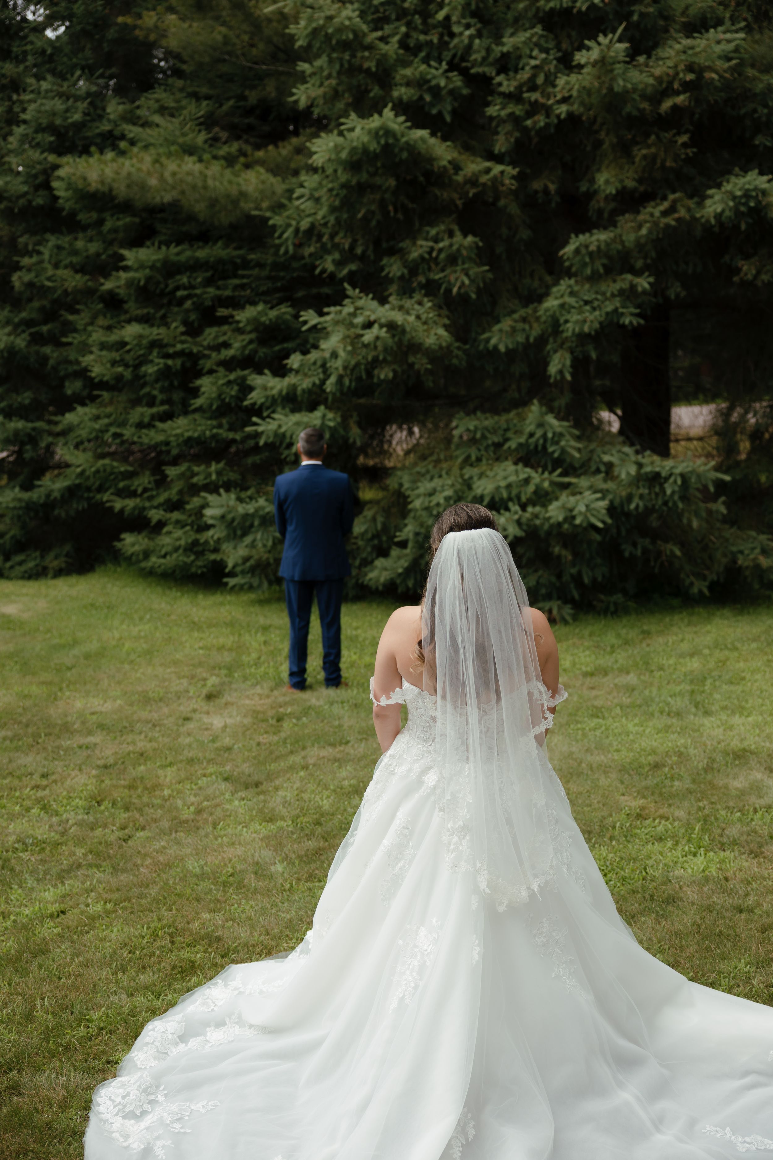 A bride in a flowing white wedding gown with a long train walks across a grassy lawn surrounded by trees.