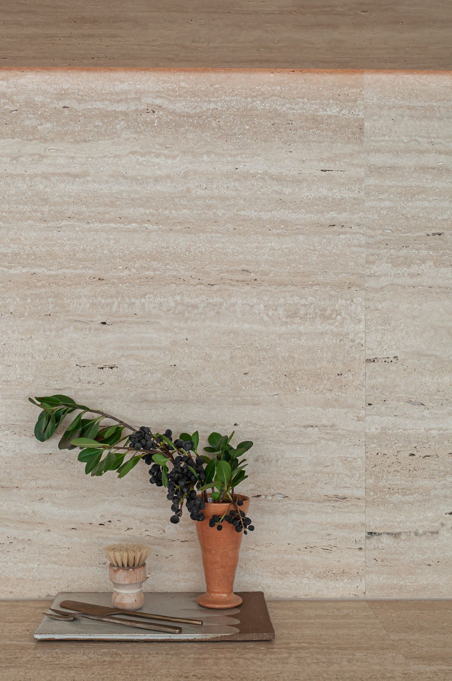 Terra cotta pottery planters with dark berries on branches against a textured beige travertine wall.