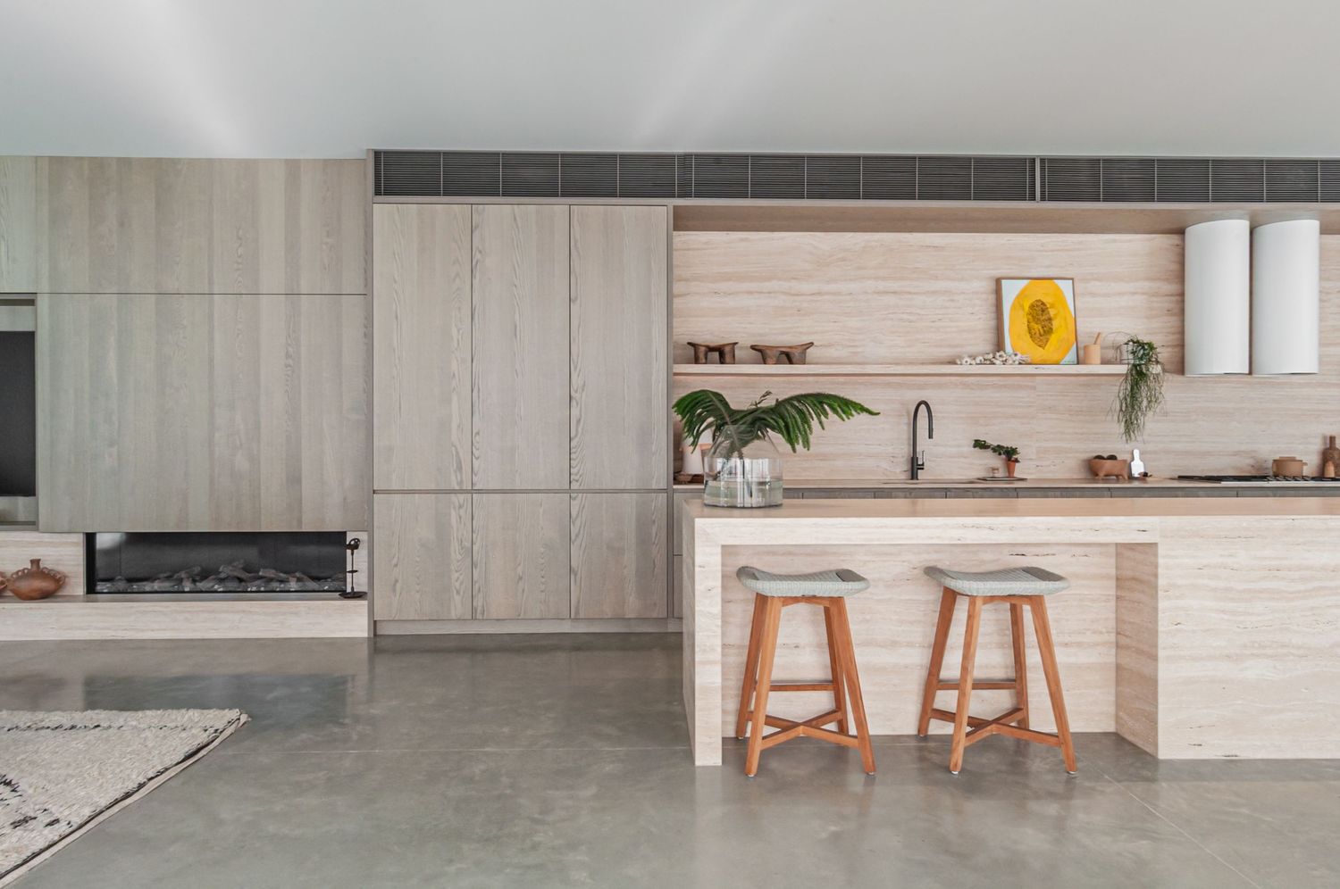 Contemporary kitchen space with light wood tones, sleek white cabinetry, and floating shelves against stone walls.