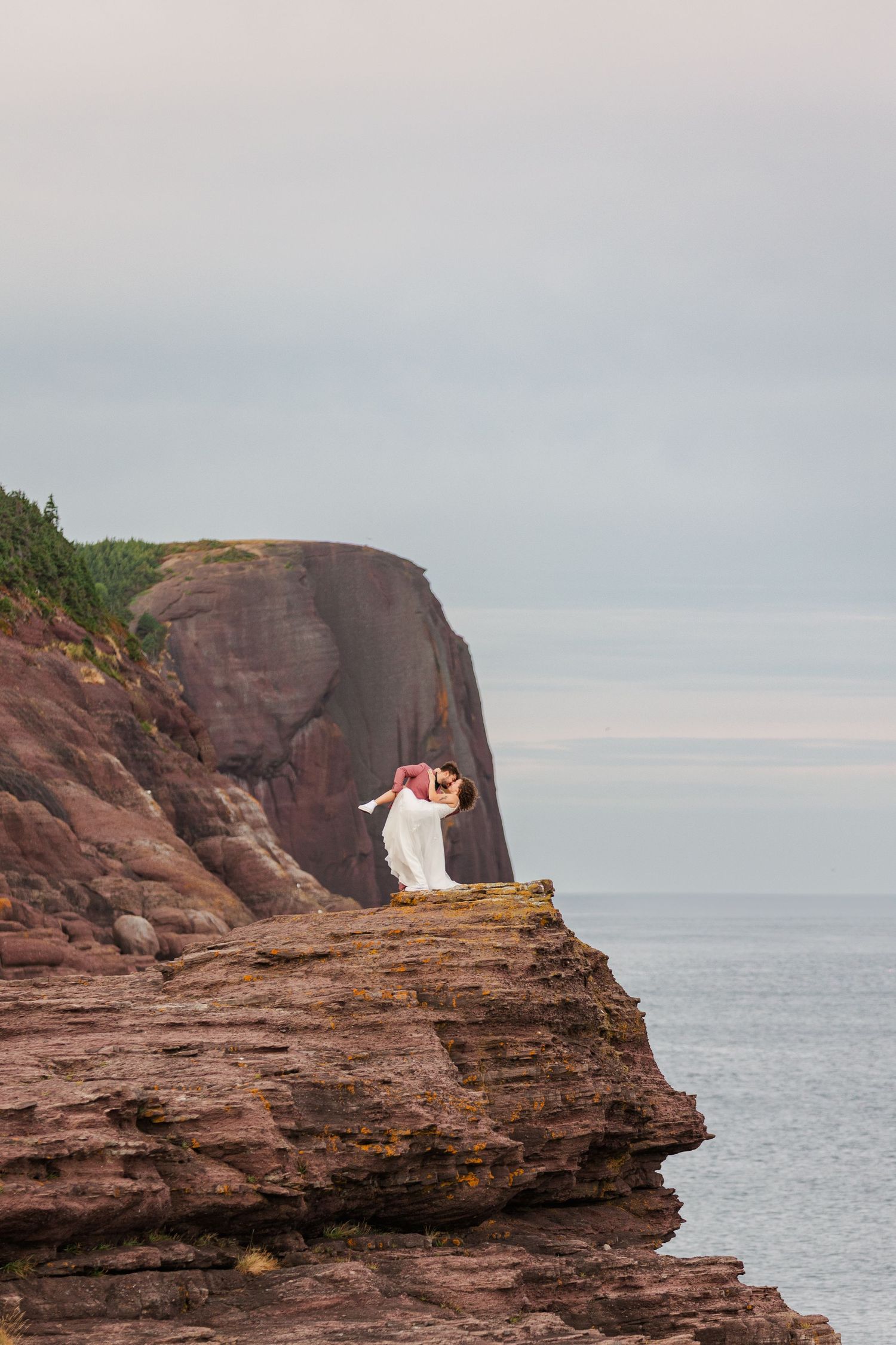 A couple in wedding attire shares an intimate moment on dramatic red sandstone cliffs overlooking the ocean at sunset.