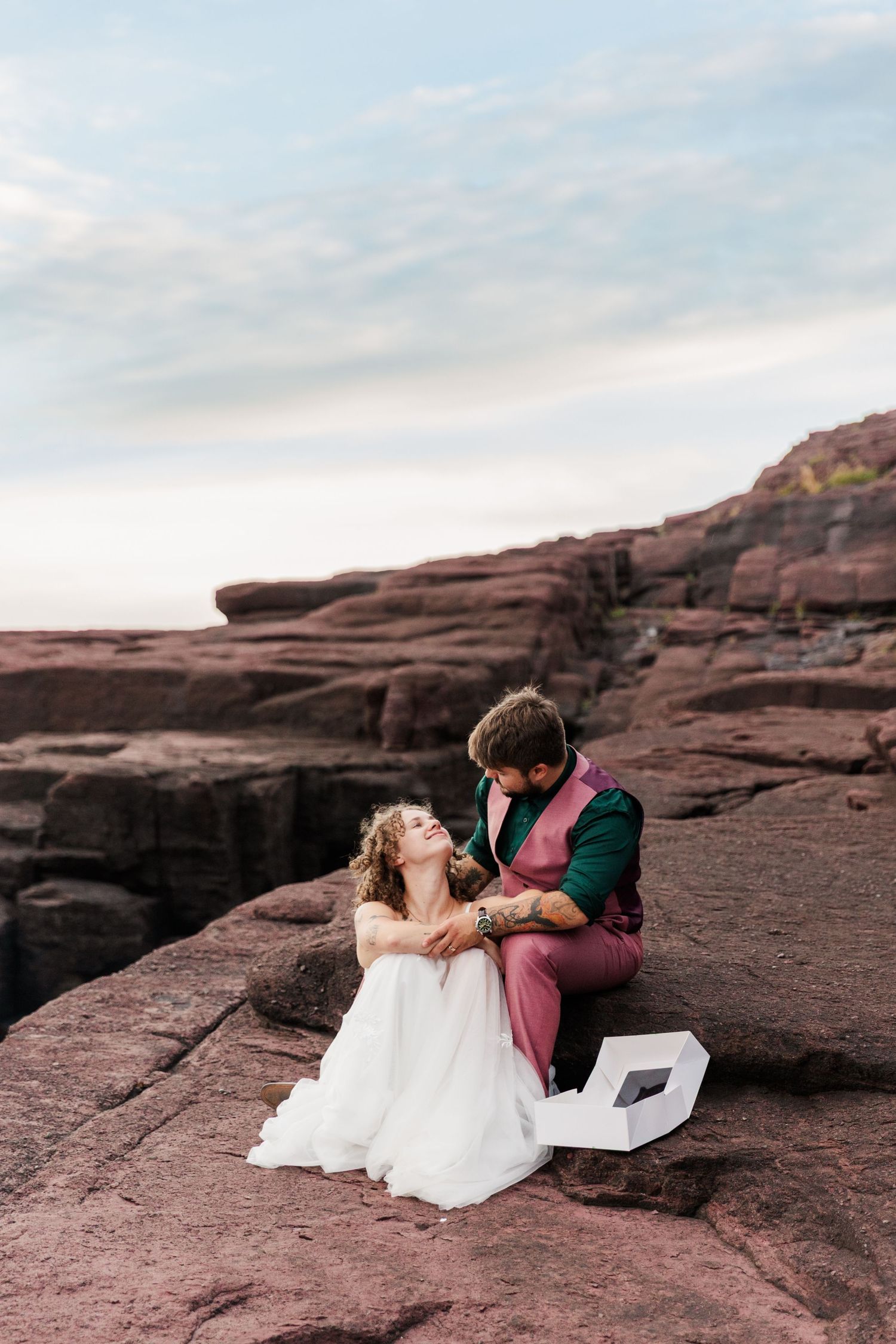 Couple sits intimately on dramatic red rock cliffs against cloudy sky during sunset photo session.